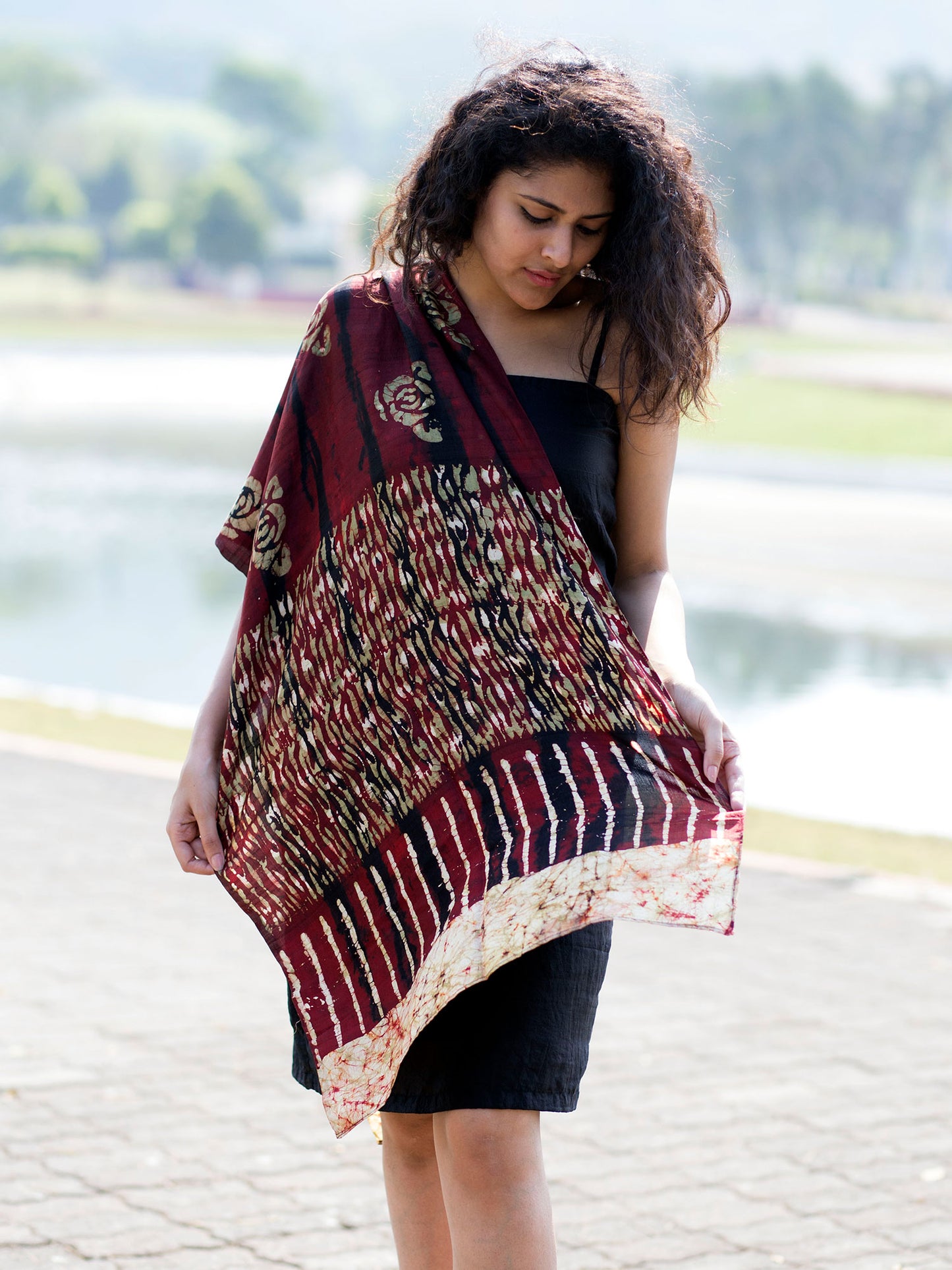 There's a young Indian woman sporting a brown silk scarf with batik prints. She is outdoor. The weather is sunny and there are trees and a lake in the background, is blurry.