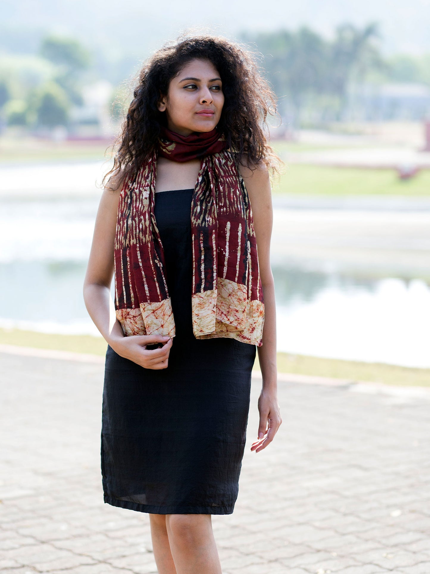 There's a young Indian woman sporting a brown silk scarf with batik prints. She is outdoor. The weather is sunny and there are trees and a lake in the background, is blurry.